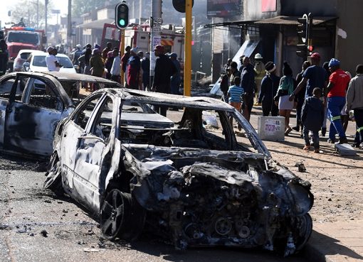 Pejalan kaki melewati mobil-mobil yang terbakar di sisi jalan di pinggiran Johannesburg pada hari Senin (2/9). (Foto: AP/Al Jazeera)
